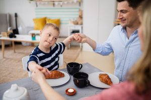 Happy down syndrome boy with parents at the table, praying before breakfast. Child with disabilities